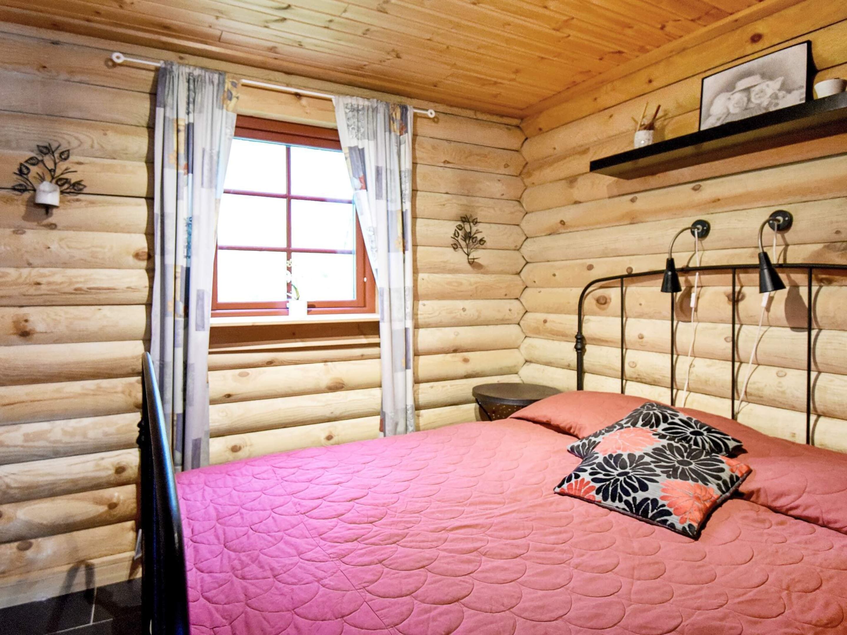 Una habitación de cabaña de troncos con una cama, una ventana con cortinas, una mesita de noche y un estante con un cuadro.