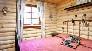 Una habitación de cabaña de troncos con una cama, una ventana con cortinas, una mesita de noche y un estante con un cuadro.