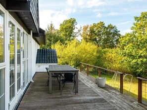 Una terraza de madera con una mesa y sillas, una valla metálica y vistas a árboles y un edificio.