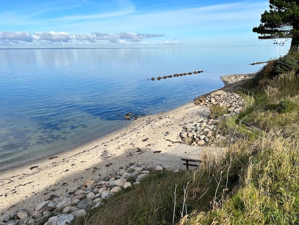 Una vista despejada de una playa junto al lago, con costa rocosa, un banco de madera y una hilera de piedras en el agua.