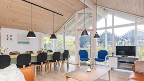 Ein modernes Wohnzimmer mit Holzboden, großen Fenstern, einem Essbereich mit Tisch und Stühlen sowie einem Fernseher auf einem Schrank.