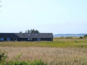 A long, dark building with multiple windows, set in a grassy field with a body of water and hills in the background. - 4 Star Holiday Home in Millinge (Millinge)