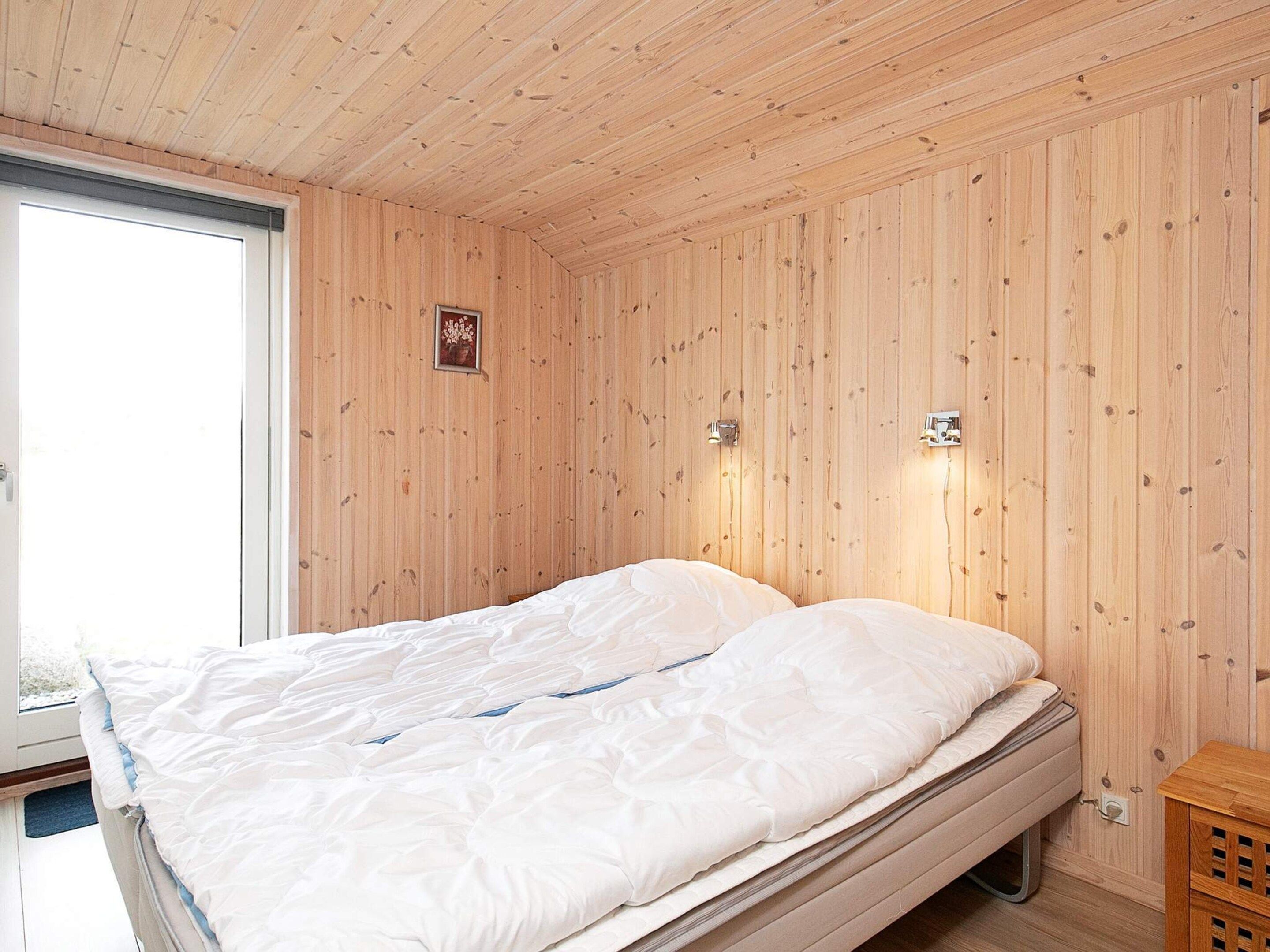 A room with wooden panel walls, a bed with white bedding, a window, and a small wooden cabinet.