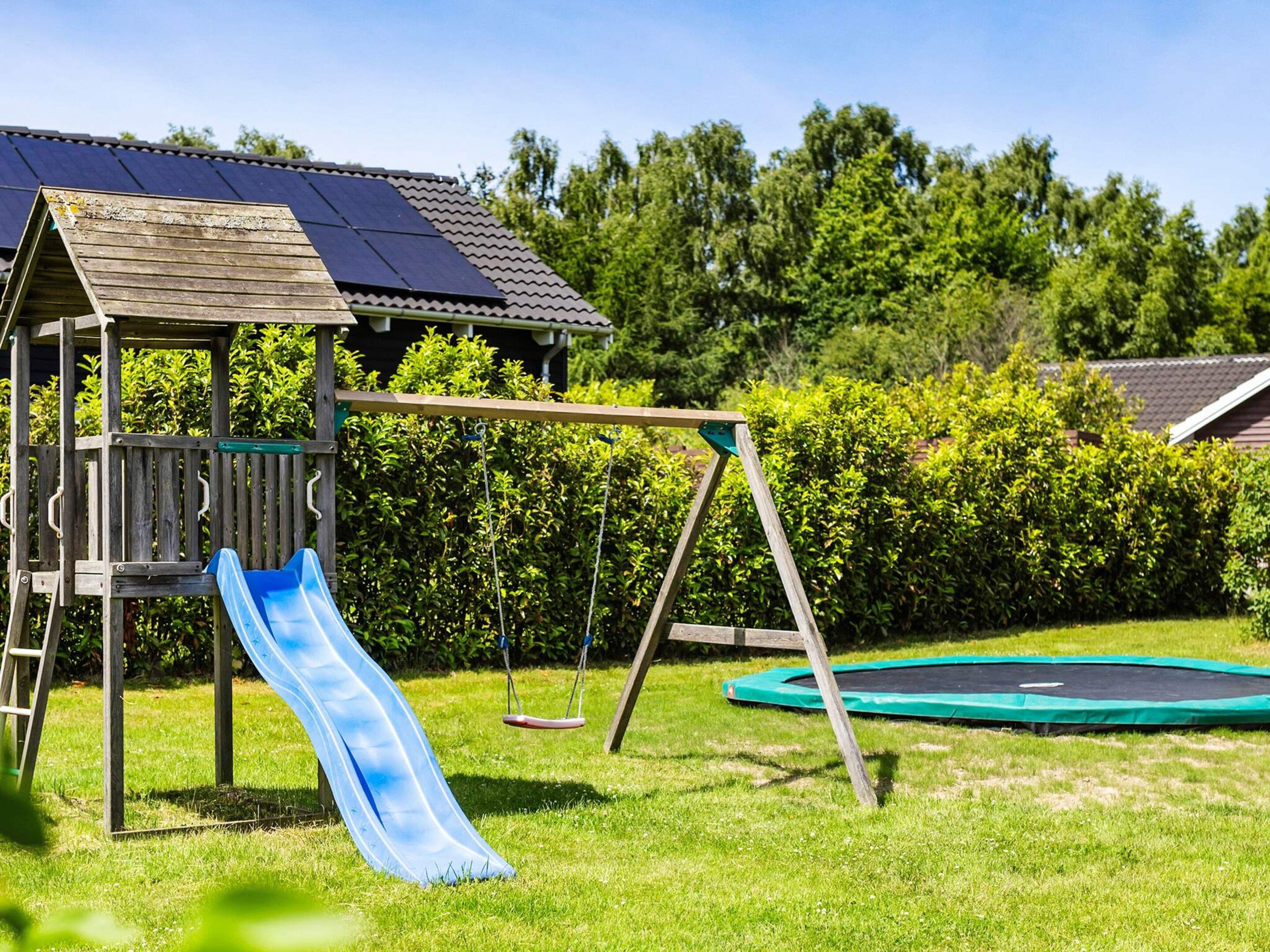 Et trælegeland med rutchebane og gyngestole, samt en trampolin og solpaneler på taget.