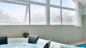 A modern bathroom with a hot tub, a potted plant, and a view of a residential area through large windows.