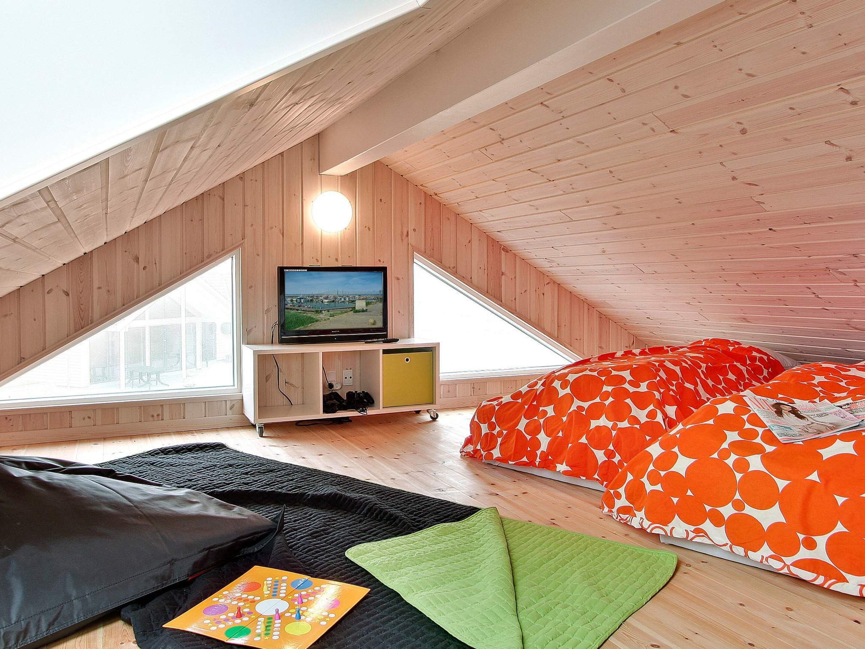 Un dormitorio tipo loft con techo inclinado, un televisor, una cama con ropa de cama naranja y blanca, y un pequeño armario de madera.