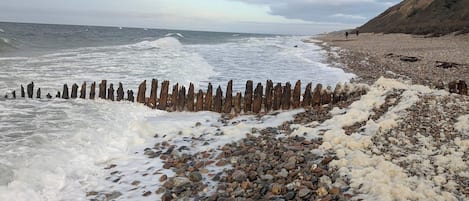 A rocky beach with waves crashing onto the shore, a wooden fence, and a hilly landscape in the background.