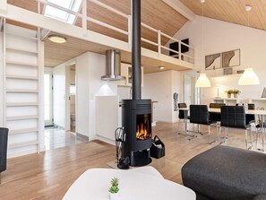 A modern living room with a wood-burning stove, a dining area with a table and chairs, and a kitchen in the background. - Modern Cottage With Spa - By Traum Ferienwohnungen (Brovst)