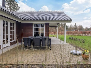 Eine überdachte Terrasse mit Gartenmöbeln, ein Ziegelweg und ein Garten mit Bäumen und Sträuchern.