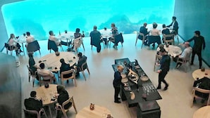 A spacious dining area with multiple round tables and chairs, a central bar area, and a large screen displaying a presentation. - 8 Person Holiday Home in Lindesnes-by Traum (Lindesnes)