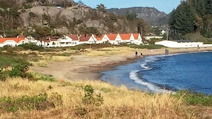 A coastal area with houses with red roofs, a sandy beach, and a rocky hillside. - 8 Person Holiday Home in Lindesnes-by Traum (Lindesnes)