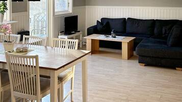 A living room with a wooden dining table, white chairs, a dark-colored sofa, and a television on a stand.