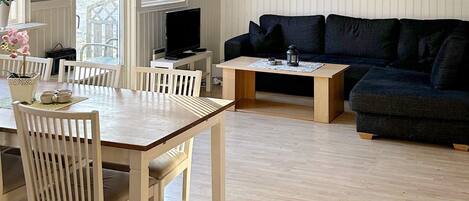 Una sala de estar con una mesa de comedor de madera, sillas blancas, un sofá oscuro y un televisor sobre un mueble.