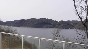 A wooden deck with a glass railing overlooking a lake with hills in the background. - 10 Person Holiday Home in Lindesnes (Lindesnes)