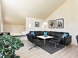 A modern living room with a sectional sofa, a coffee table, and a dining table. There are framed pictures on the wall, a ceiling fan, and a potted plant.