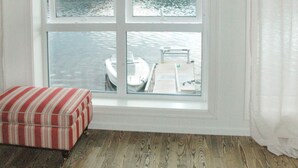 Een kamer met een houten vloer, een gestreept voetenbankje, een raam met uitzicht op een steiger en boten, en een wit gordijn.