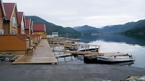 Een rij houten gebouwen met rode daken aan een houten steiger die uitloopt in een rustig meer, omringd door bergen.