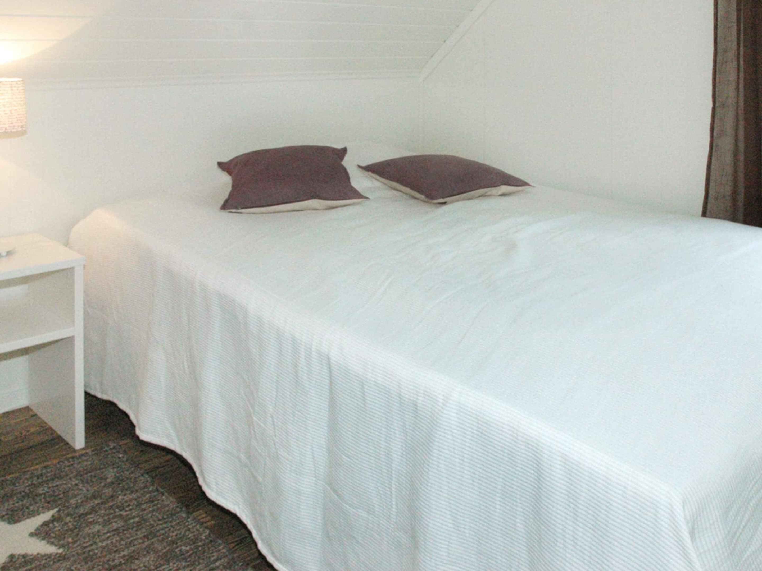 Una cama individual con colcha blanca y dos almohadas, una mesita de noche blanca y un tapete estampado sobre piso de madera.