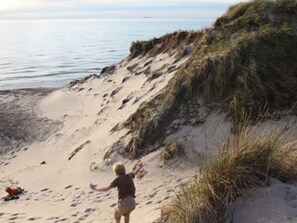 Una playa con una persona de pie sobre la arena, una duna y el océano al fondo.
