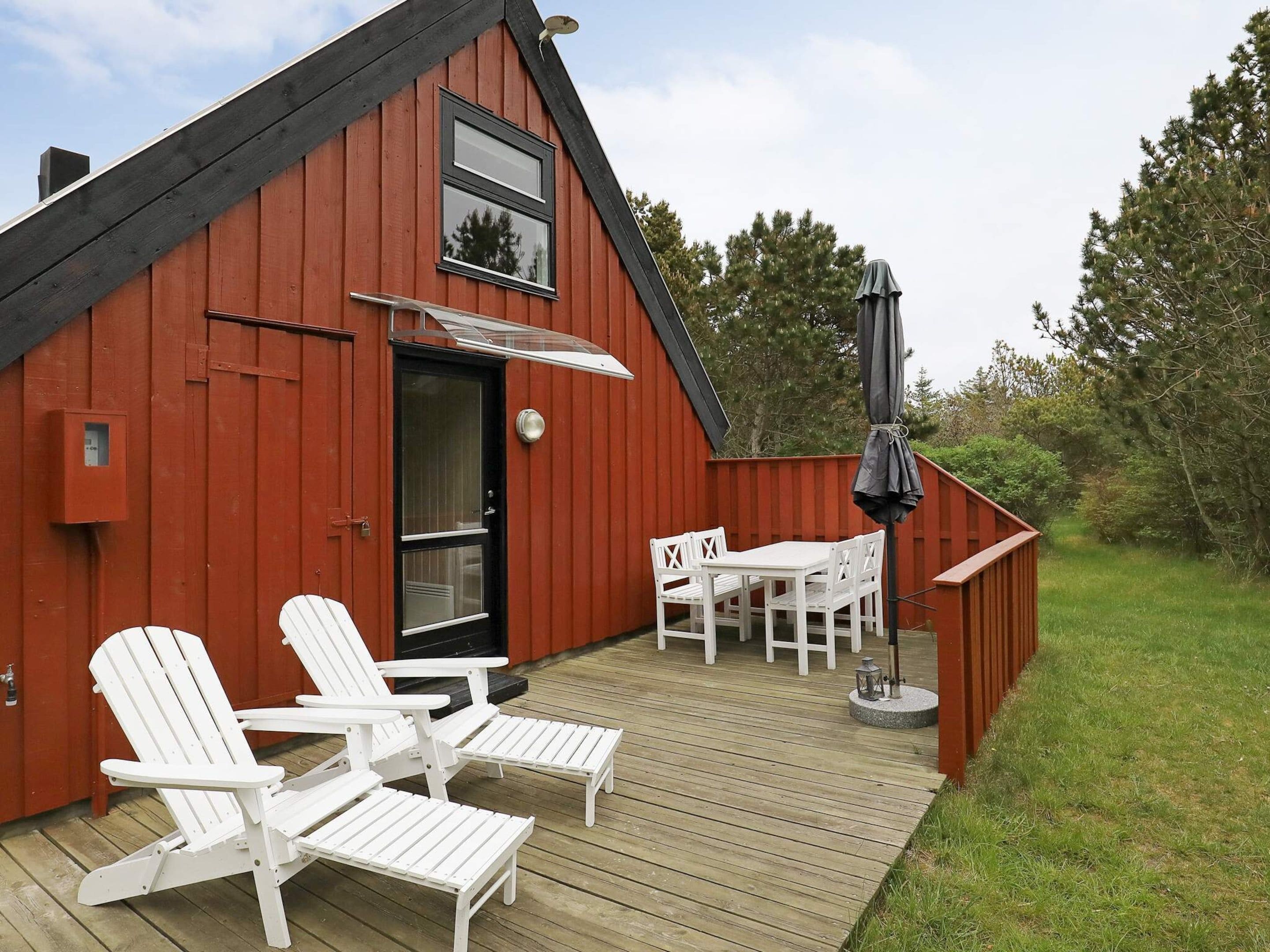 Een rood houten gebouw met een zwart dak, een deur en een raam. Er is een terras met witte stoelen en een tafel, en een zwarte parasol.