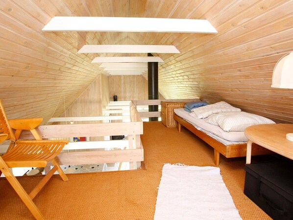 Una habitación con techo inclinado, paredes de madera, una cama con almohadas, una silla, una mesita y una alfombra blanca.