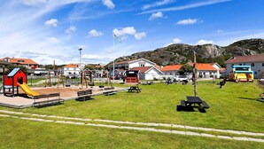 Ein Spielplatz mit Rutsche, Schaukeln und Picknicktischen in einem Park, im Hintergrund Wohnhäuser.