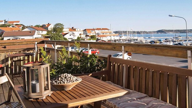 Eine Holzterrasse mit Tisch und Bank, die auf einen Yachthafen mit Booten und Häusern im Hintergrund blickt.