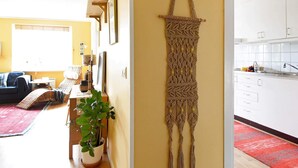 Ein Wohnzimmer mit einer Couch, einem Couchtisch und einer Topfpflanze. An der Wand hängt ein Wandbehang aus Makramee.