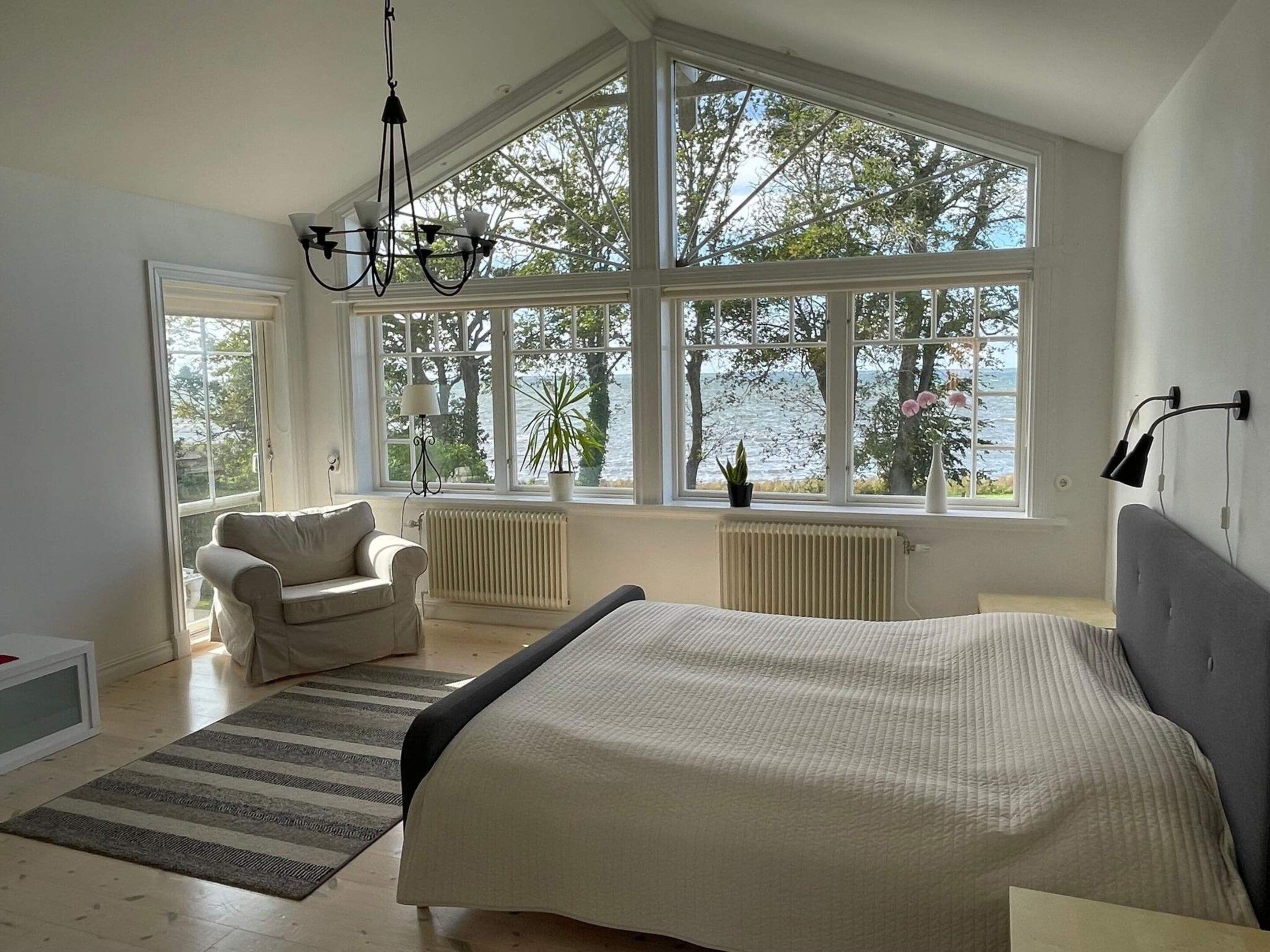A bedroom with a large bed, a chair, a view of the sea through large windows, and a chandelier.