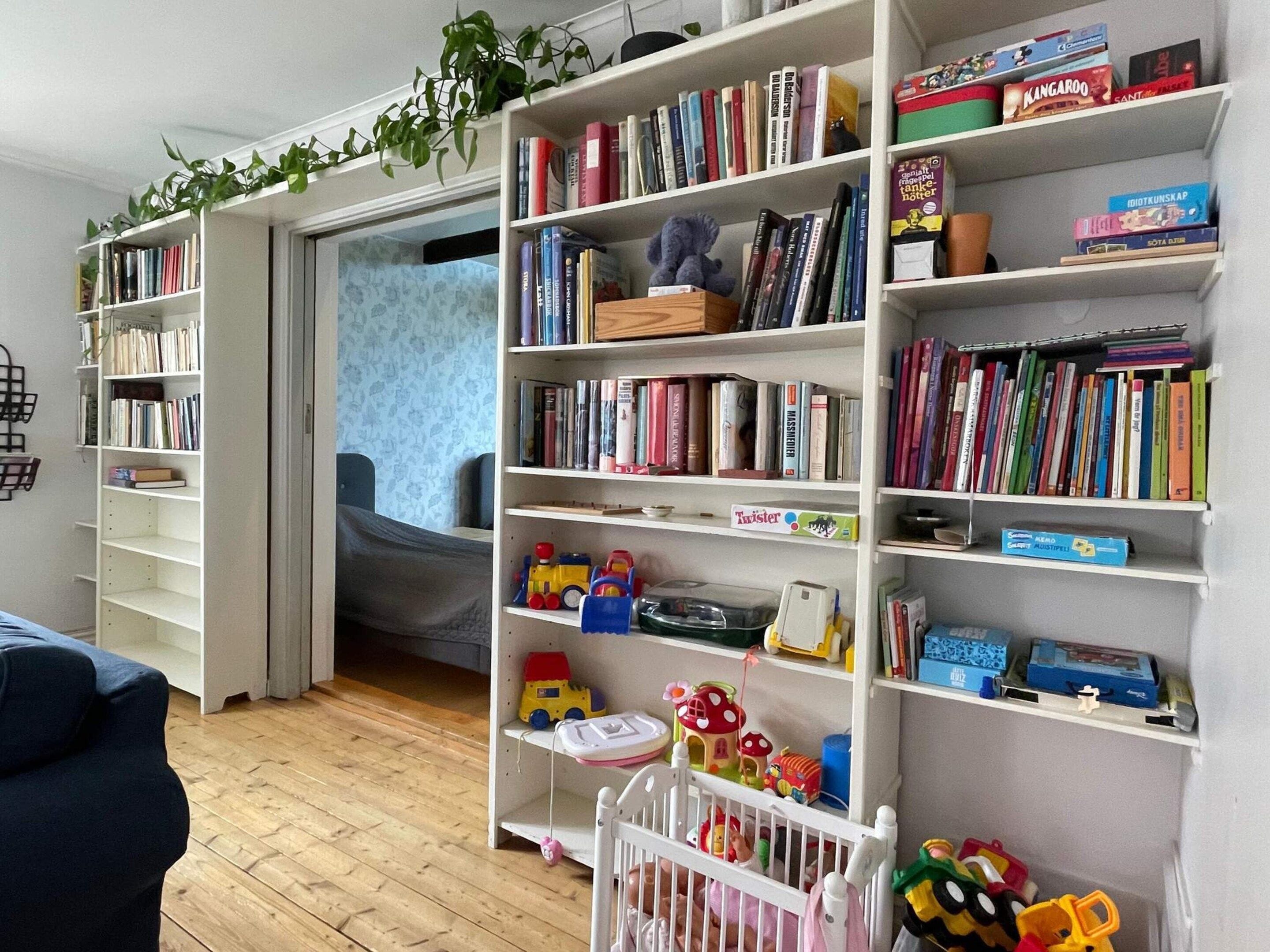 A room with a bookshelf filled with books and toys, a crib, and a blue couch.
