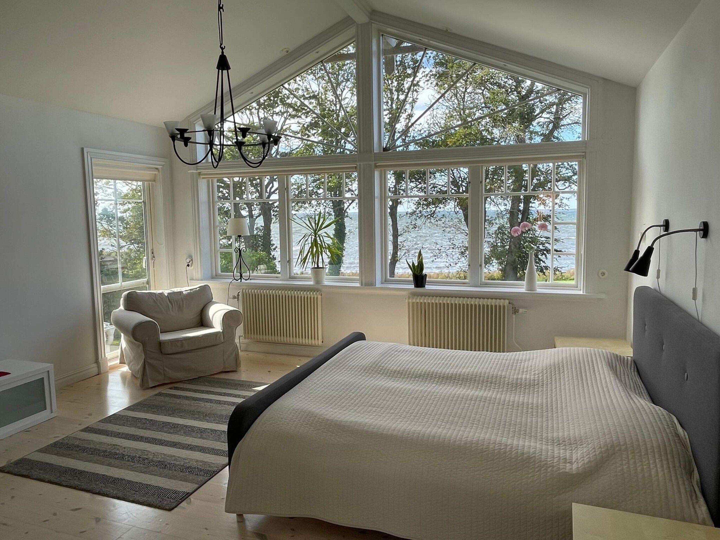 A bedroom with a large bed, a chair, a view of the sea, and a window with a plant.