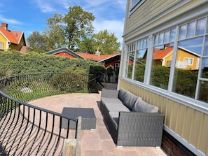 A balcony with a curved metal railing, a grey cushioned seating area, and a view of neighboring houses and trees. - Holiday Home in Mörbylånga (Morbylanga)