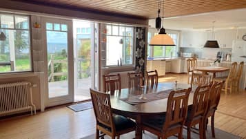 A spacious dining area with a wooden table and chairs, a hanging light fixture, and large sliding glass doors leading to a balcony with a view of the sea.