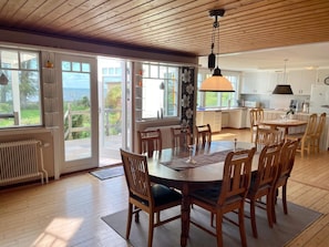 Uma ampla área de jantar com mesa e cadeiras de madeira, luminária pendente e grandes portas de vidro deslizantes que dão para uma varanda com vista para o mar.