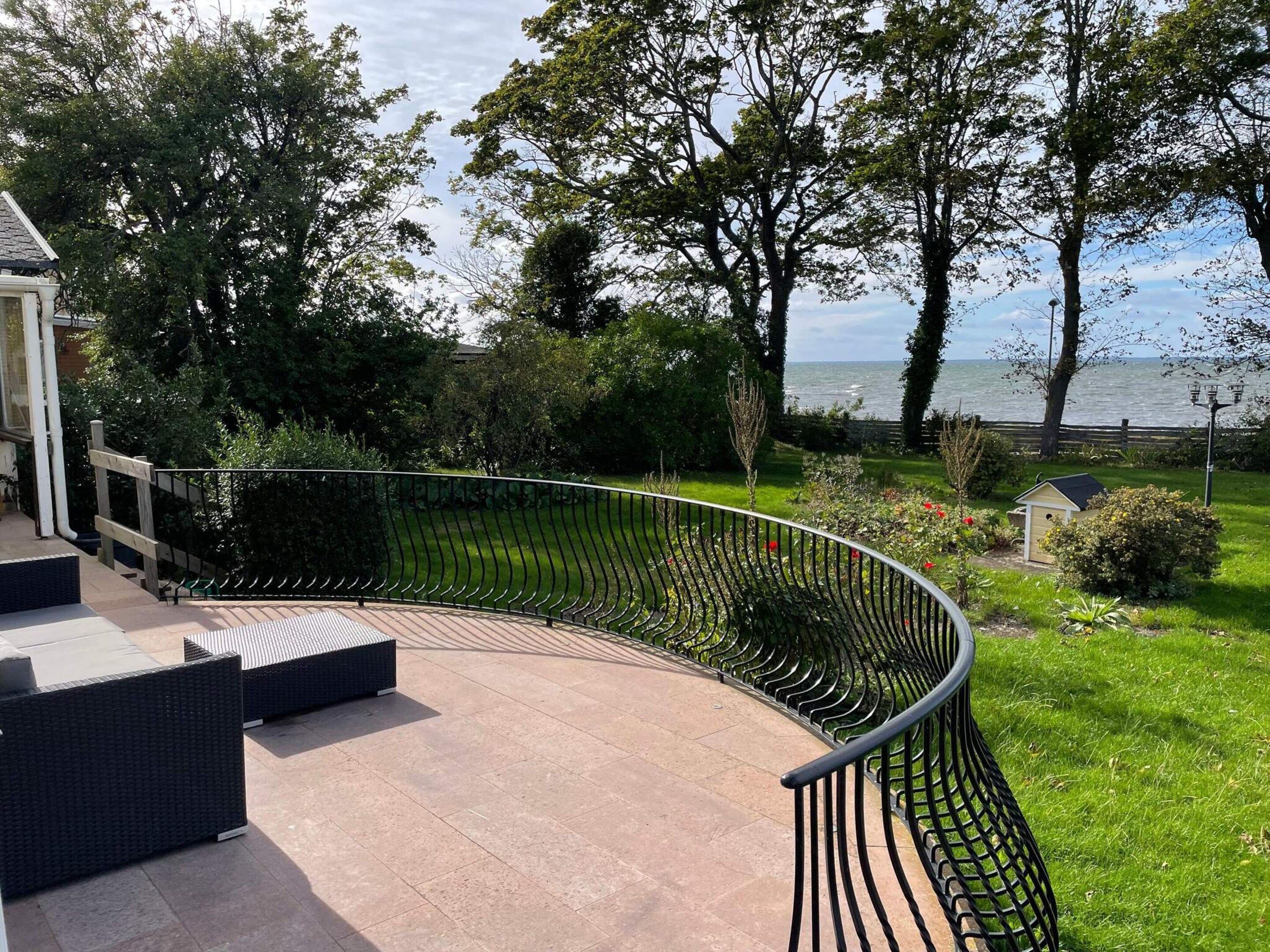 A curved outdoor patio with a metal railing, a small garden area with a shed, and a view of the sea.