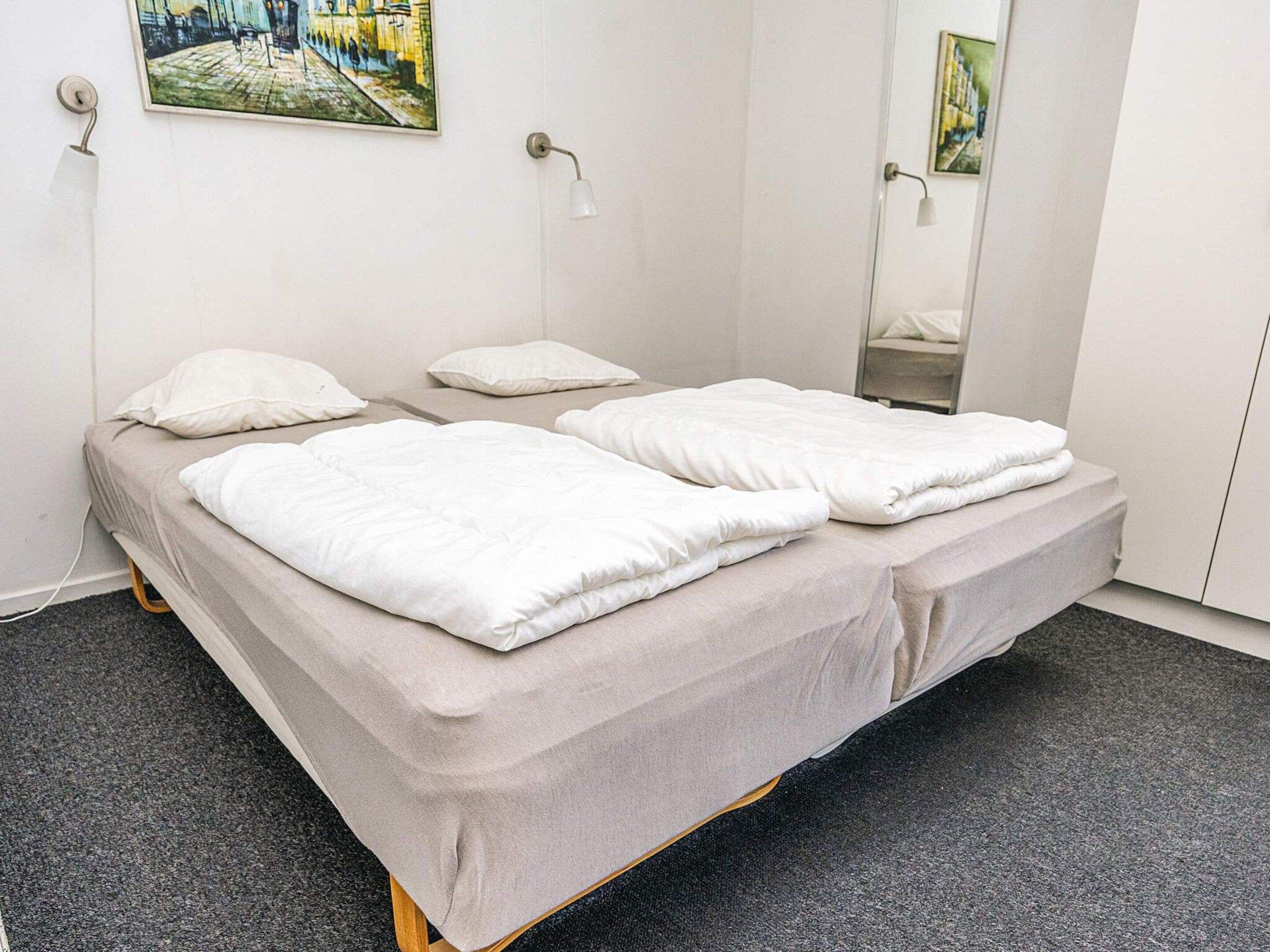 A single-bed room with a grey mattress, white pillows, a wooden frame, a grey carpet, and two wall-mounted lamps.