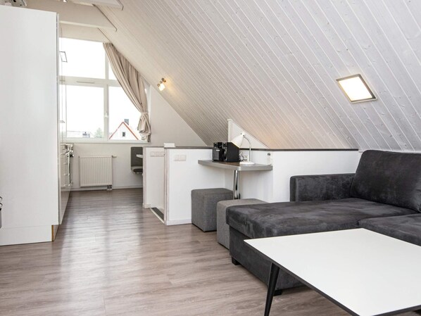 A modern loft apartment with a sloped ceiling, a kitchenette, a gray sofa, a white coffee table, and a view of a house through a large window.