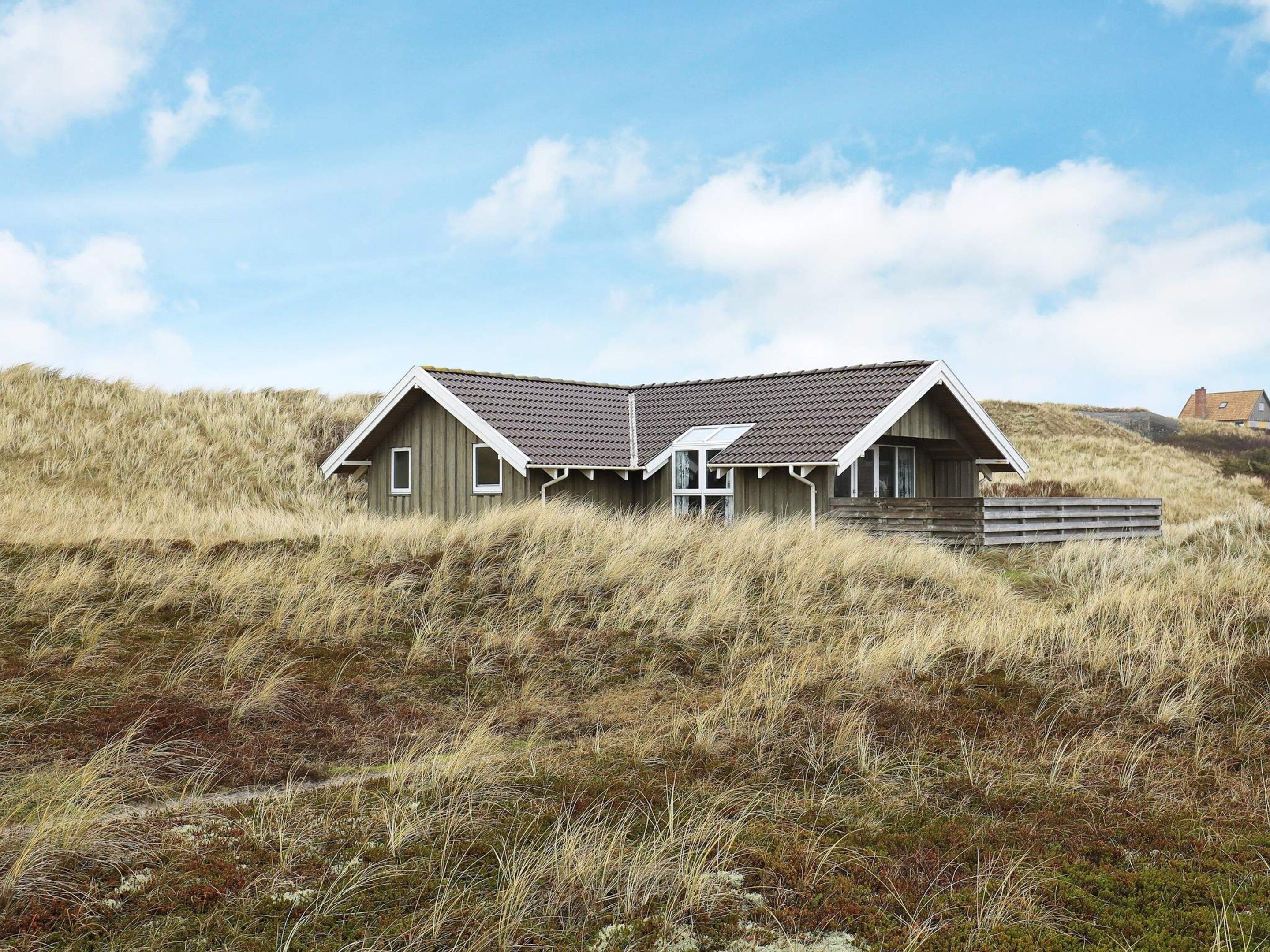A single-story wooden house with a sloped roof, surrounded by tall grass and dunes.