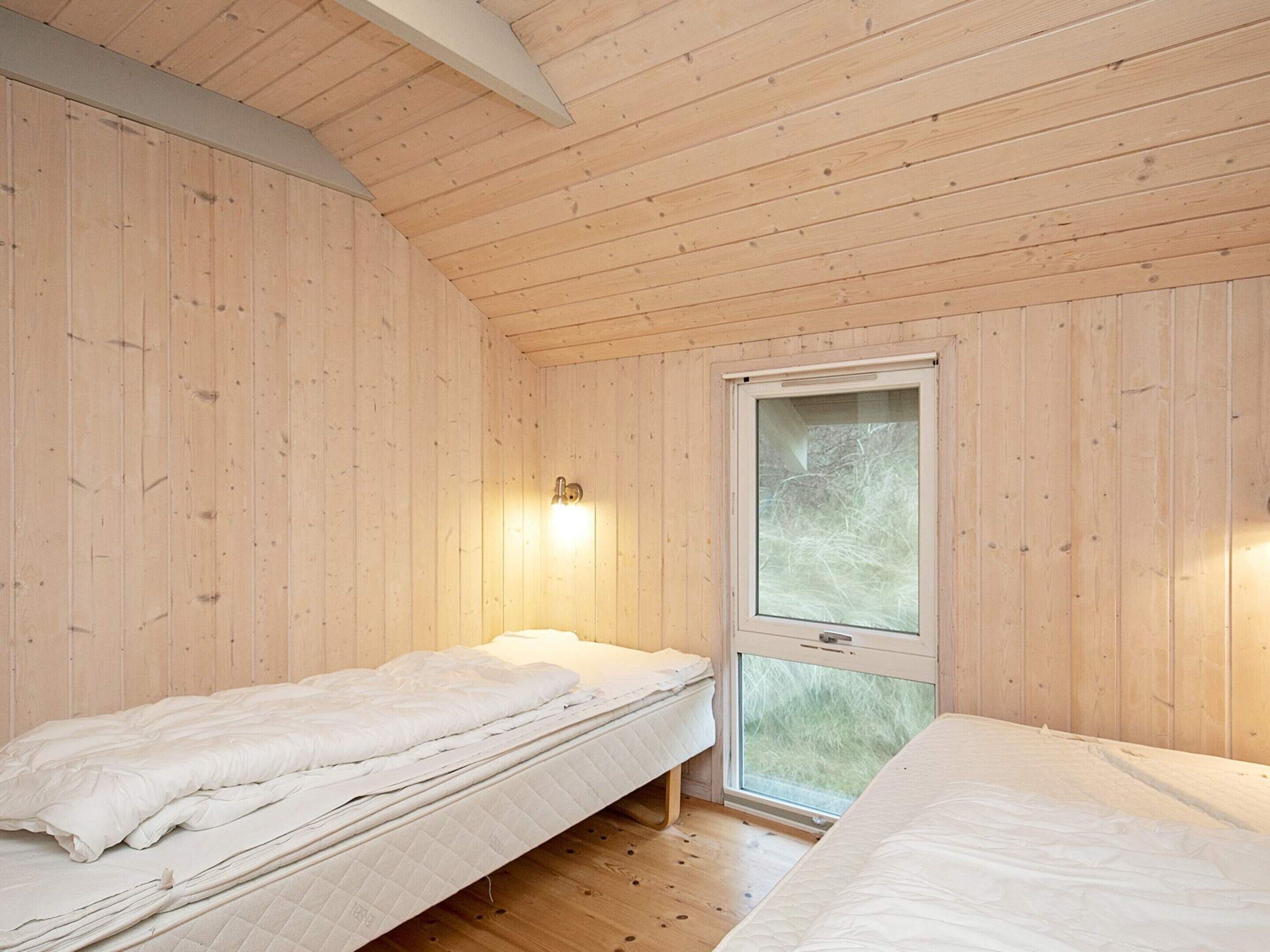 A room with wooden walls and floors, a single bed, a window with a view, and a wall-mounted light fixture.