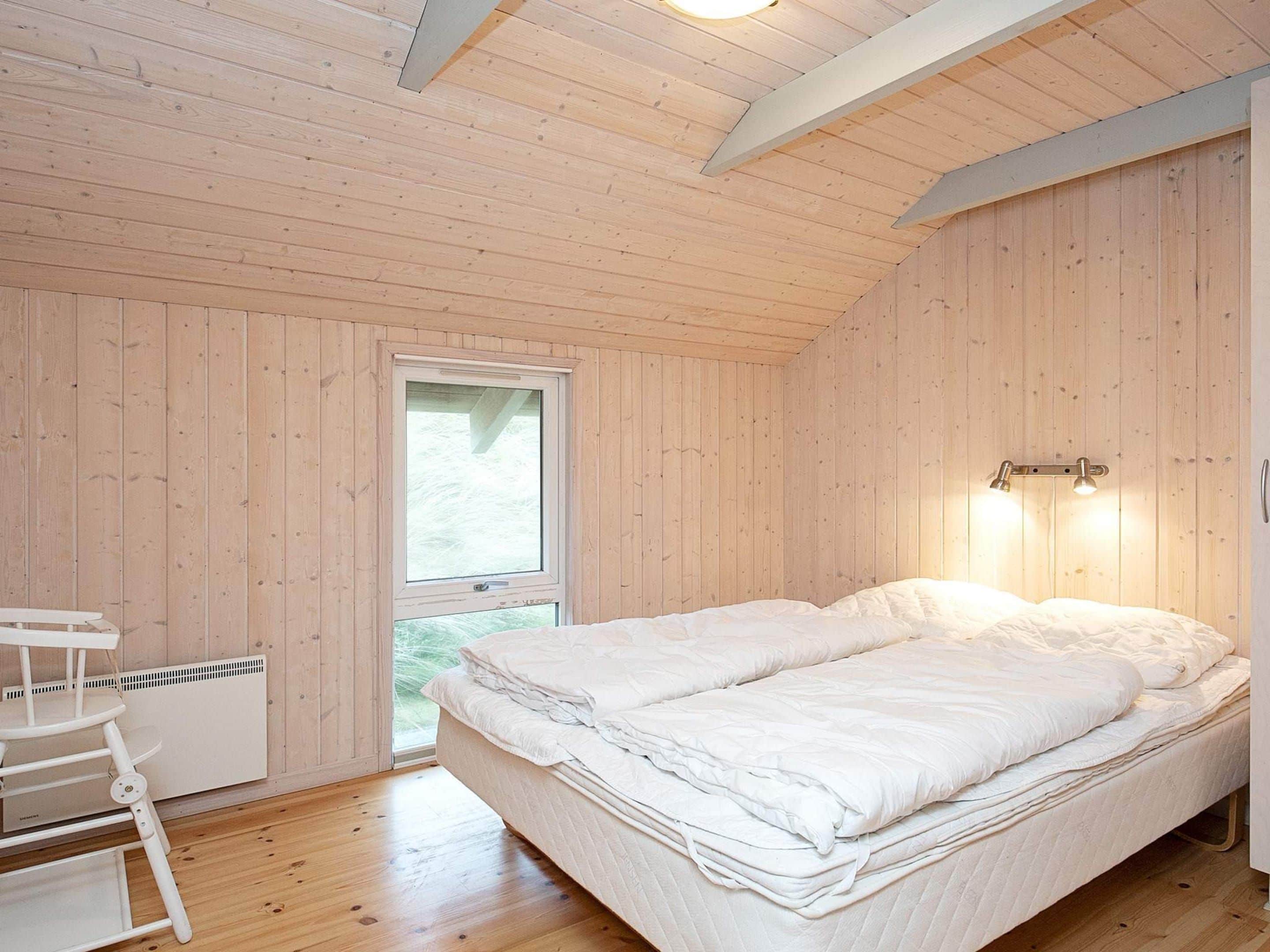 A room with wooden walls, a bed with white bedding, a window, and a desk with a chair.