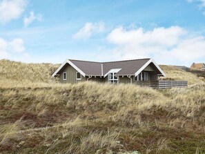 A wooden house with a sloped roof, surrounded by tall grass on a hill.