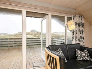 A room with a black leather sofa, patterned cushions, a wooden bench, and a view of the beach and dunes through large glass doors.