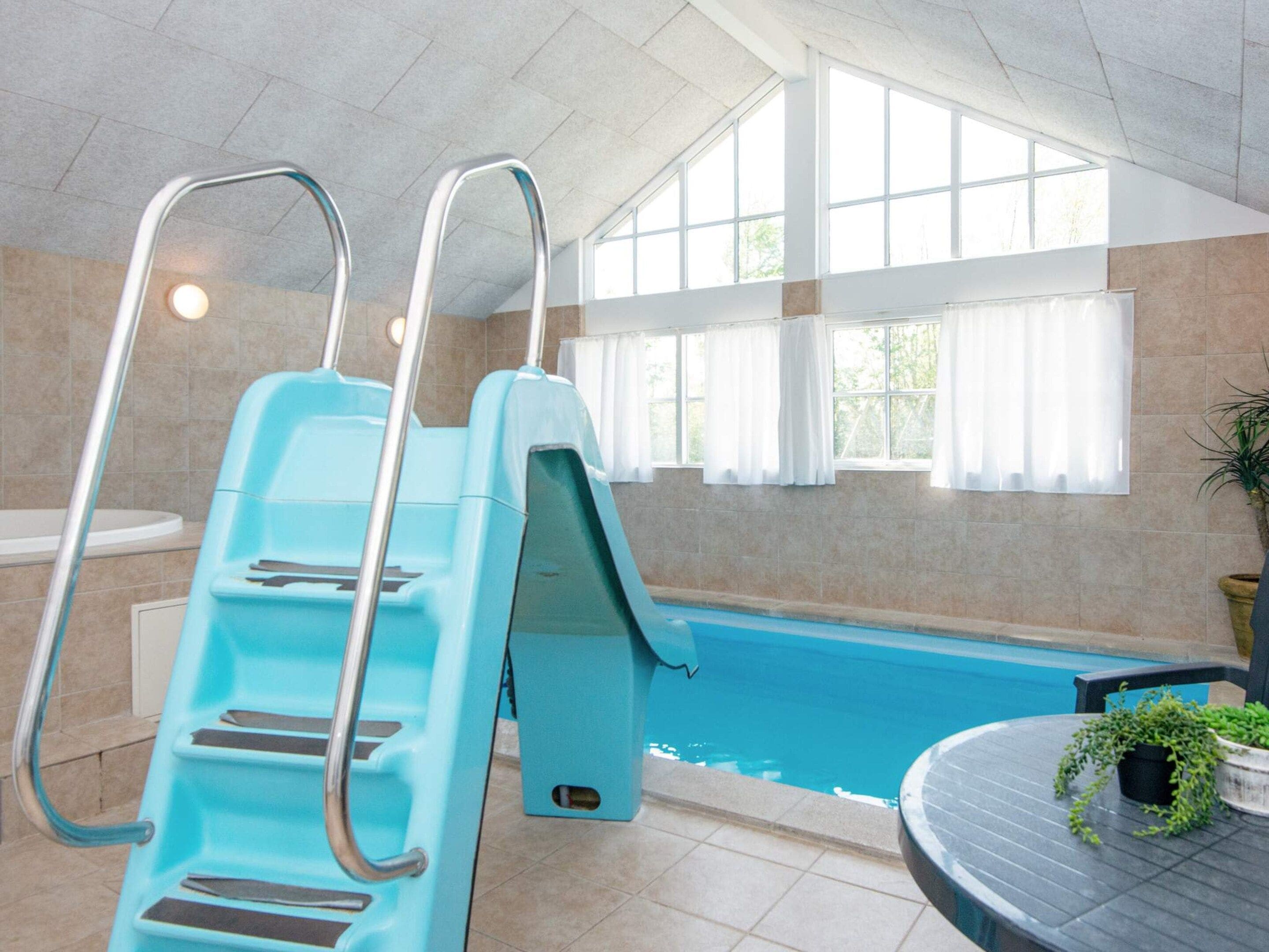 An indoor swimming pool with a blue slide, a sunlit room with large windows, and a round table with a potted plant.