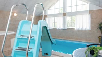An indoor swimming pool with a blue slide, a sunlit room with large windows, and a round table with a potted plant.