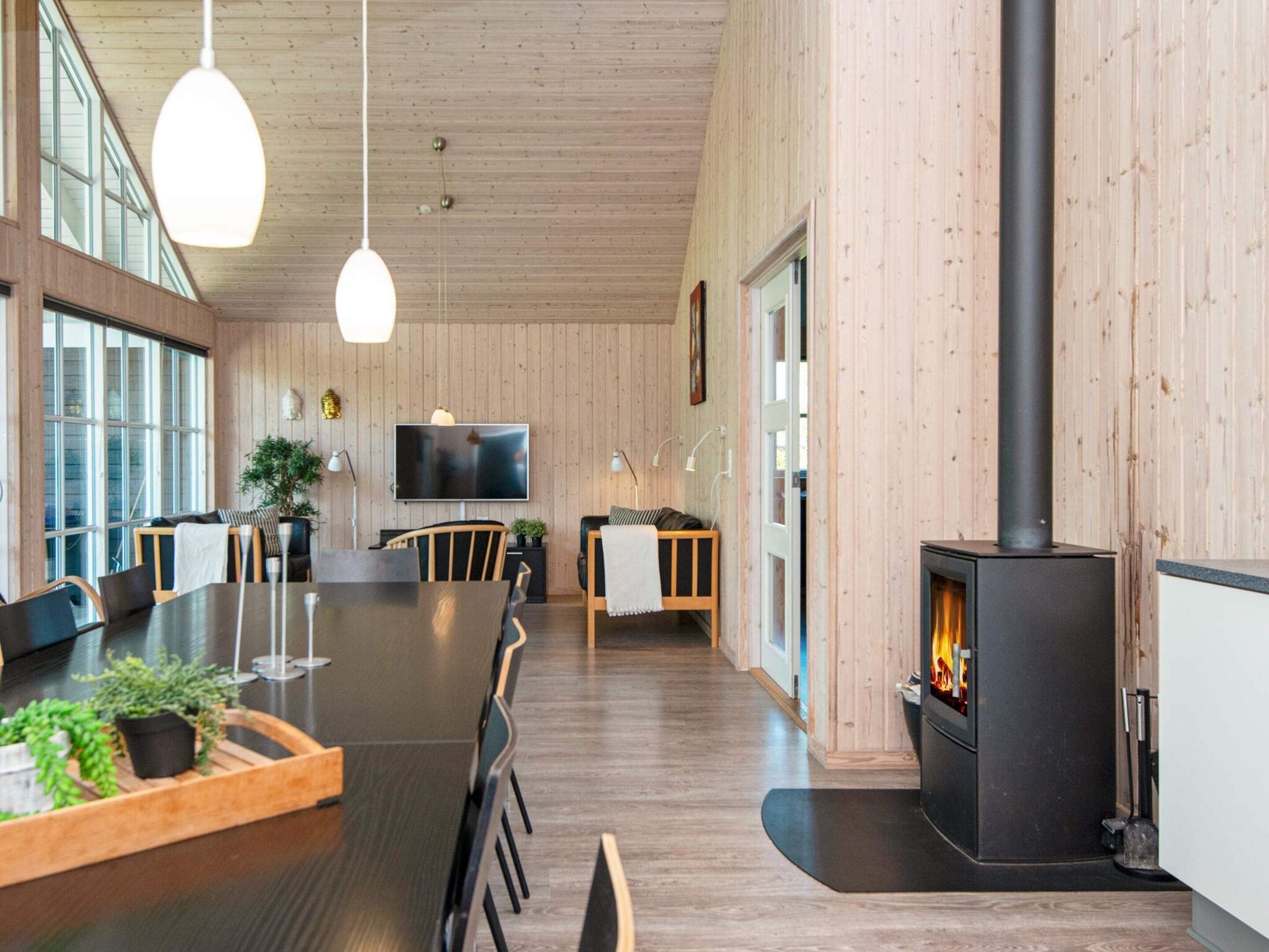 A modern interior with a long dining table, chairs, a TV, and a wood-burning stove.