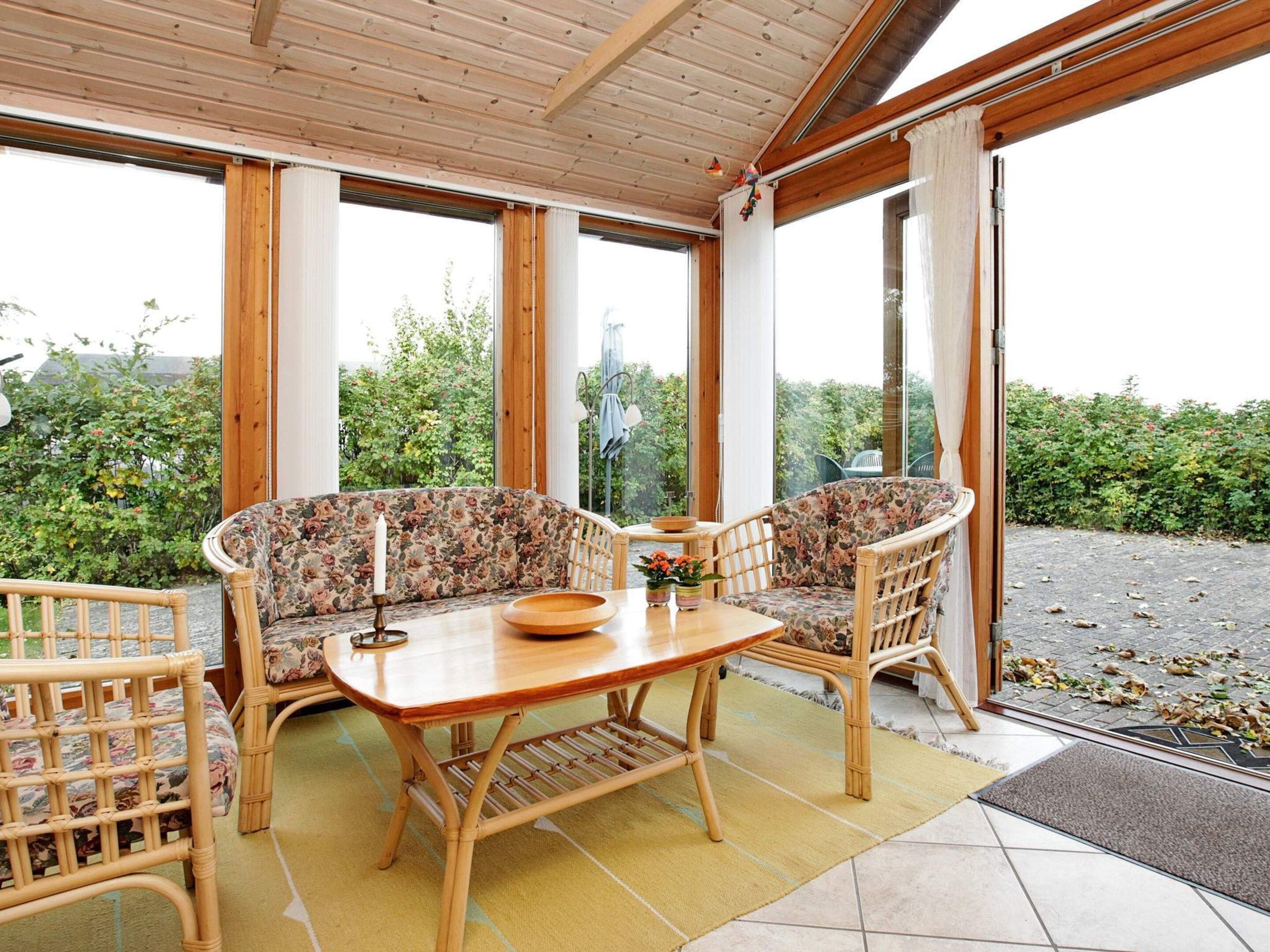 A conservatory with a wooden table and two wicker chairs, large windows, and a view of greenery.