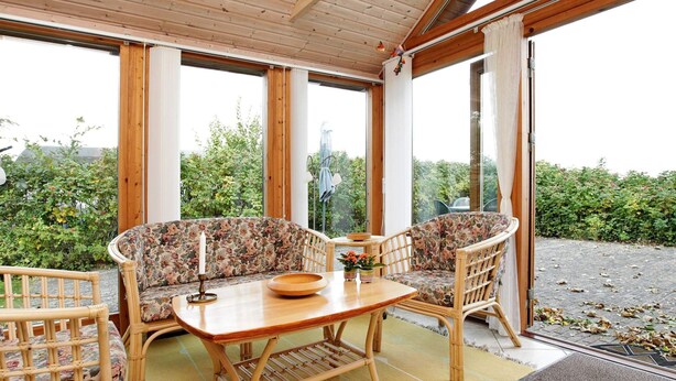 Une véranda avec une table en bois et deux chaises en osier, de grandes fenêtres et une vue sur la verdure.