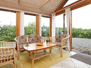 Une véranda avec une table en bois et deux chaises en osier, de grandes fenêtres et une vue sur la verdure.