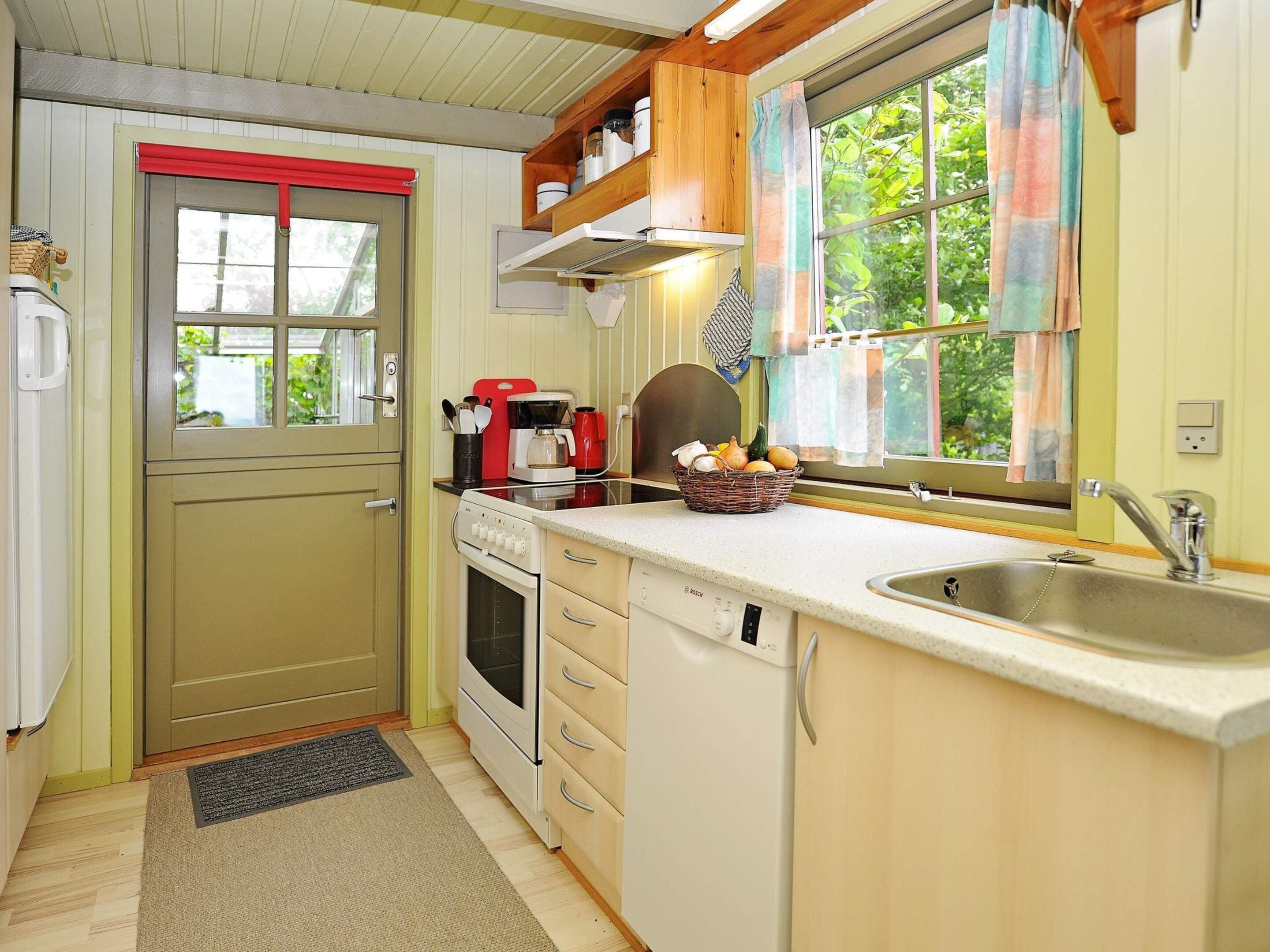 Cocina compacta con estufa, horno y pileta, muebles de madera y un ventanal con cortinas.