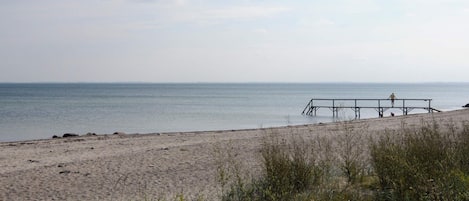 A beach with a wooden pier extending into the water, a person standing on it, and a clear sky.